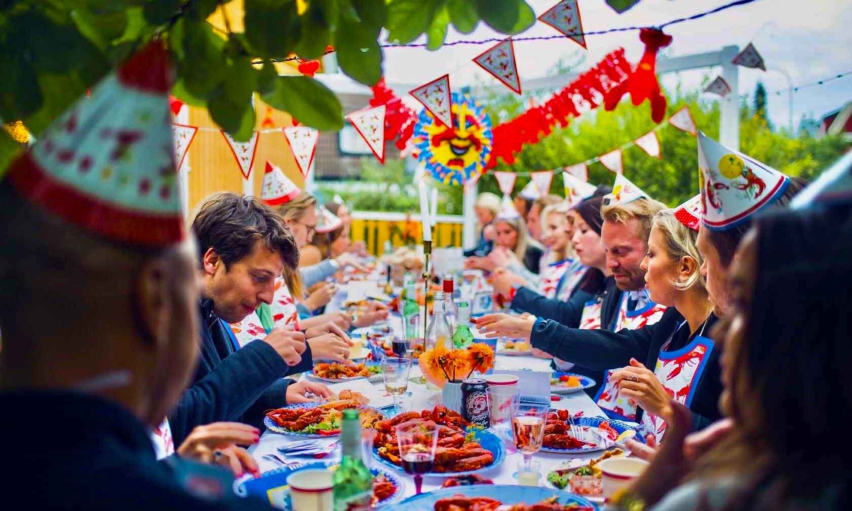 Friends in paper crayfish hats sharing a Kräftskiva table by the water